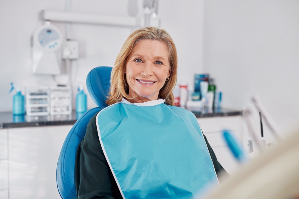 Old beautiful lady smiling while seated on dental chair wearing a blue bib for dental implant.